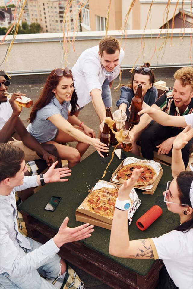 Friends on a rooftop clinking bottles over pizza, mid-laugh