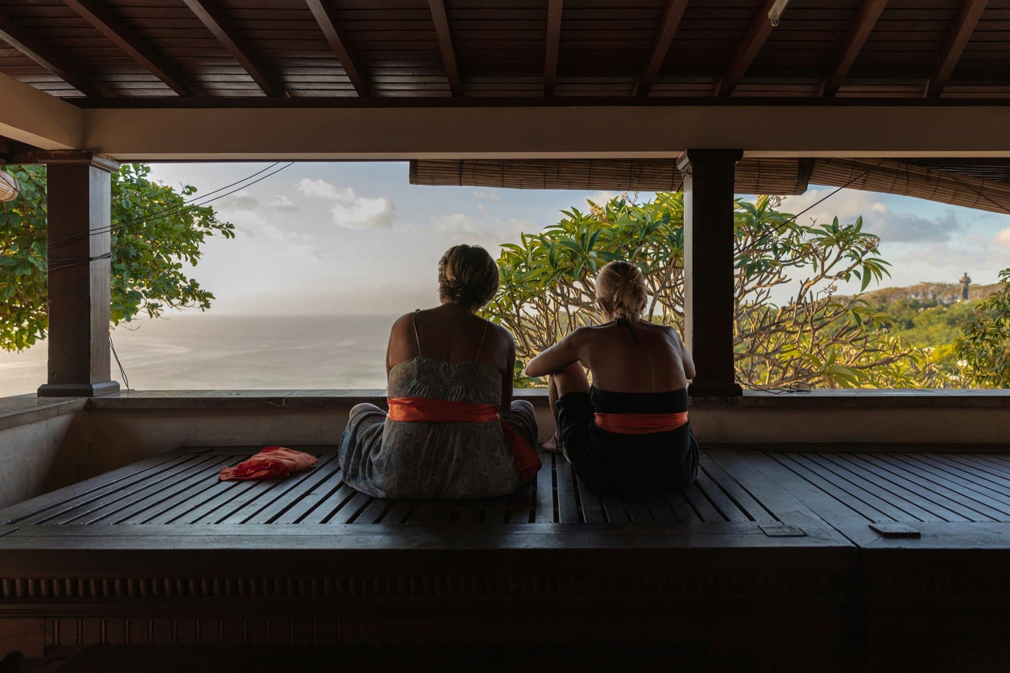 Two people sitting in the shade of a covered veranda, looking out over trees to the sea